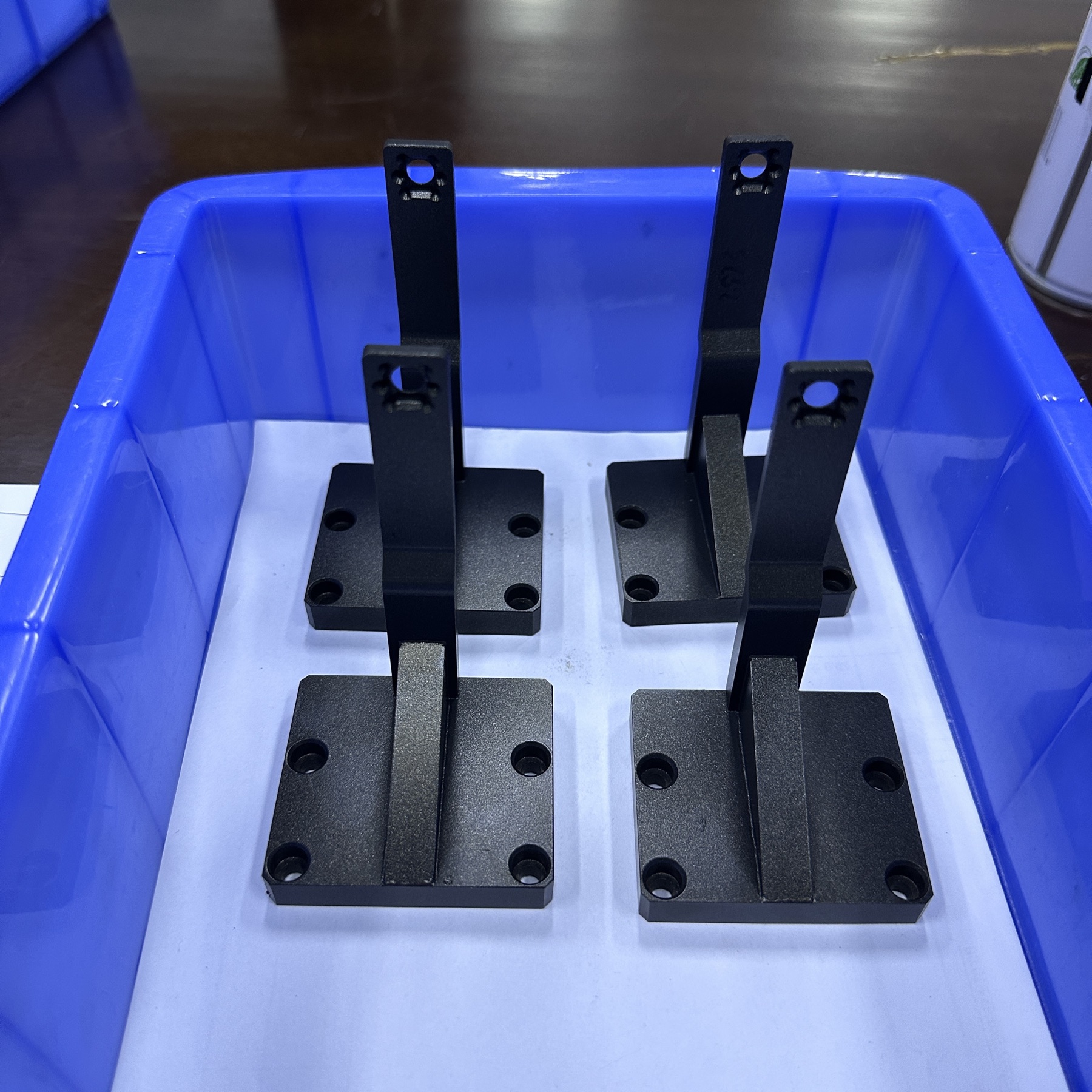 Four black CNC-machined mounting brackets with base mounting holes and upright connection arms in a blue factory tray